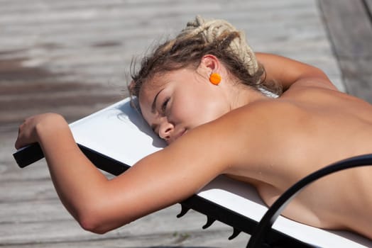 A nude woman with dreadlocks reclines on a lounge chair, enjoying the sun and a moment of relaxation outdoors.