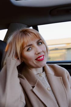 Shoulder length portrait of one young beautiful Caucasian blonde woman looking at camera with smile and sitting with head resting on hand on back seat of car while driving one spring day, side view close up. Concept time road trip, travel time.
