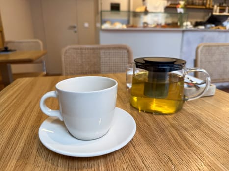 Glass teapot with herbal infusion and white ceramic cup on cafe table. Relaxation and modern tea culture.