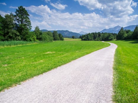 Landscape view in the Bavarian foothills of the Alps with a gravel path running into the distance in summer with a view of the first foothills of the High Alps in the background.
