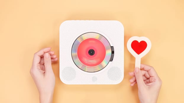 A vintage white retro styled portable compact disc player sits next to a vibrant red heart-shaped object, beautifully isolated on a soft peach background, creating a nostalgic and romantic atmosphere.