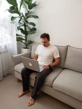 Middle-aged barefoot man in knit pants and white t-shirt using laptop on modern gray sofa in minimalist living room with fiddle leaf fig. Ideal for remote work, home office, freelance, and digital lifestyle