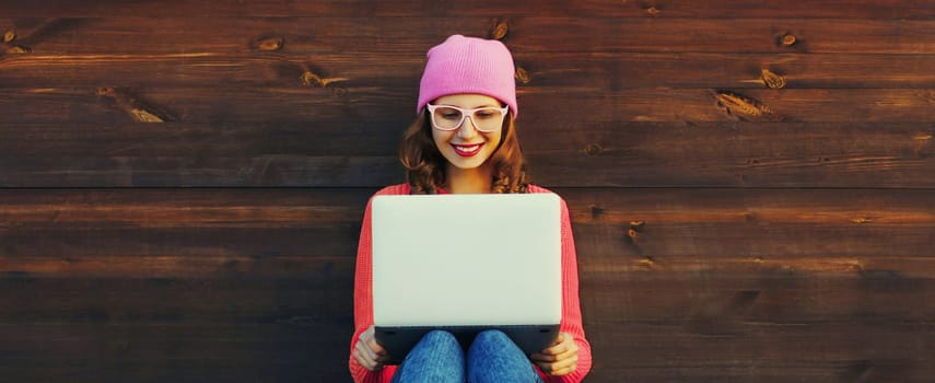 Happy modern young woman working on a laptop, freelancer girl looking at computer with glasses sitting in urban setting on wooden wall background