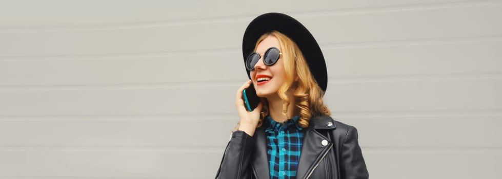 Beautiful happy smiling young woman calling on phone, stylish girl talking on the smartphone in black hat, jacket on gray background