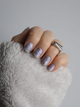 Stylish close-up of white crelly nail polish with blue shimmer, aurora flakes adorned with black foil pieces featuring silver ring and sleeve of light gray fleece sweater in neutral palette