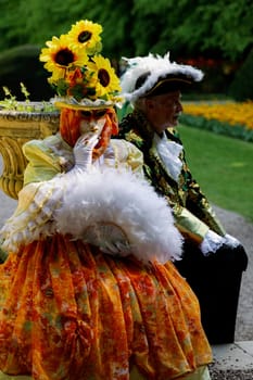 Dilbeek, Grand Bigard Castle, Belgium - April 27, 2025: Full length portrait of one couple, woman in orange dress with sunflowers on hat and man in Venetian costumes, sitting and relaxing alone in the park during the final weekend of Floralia Brussels festival, side view close up.