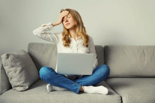 Sad upset overworked young woman suffering from headache, stress, migraine while working with laptop sitting on the couch in living room at home