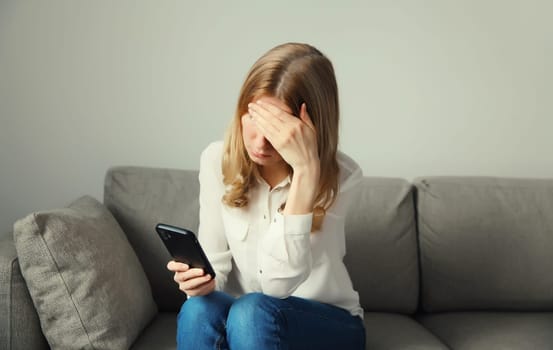 Sad upset crying young woman holding phone suffering from stress after reading emotional message sitting on the couch in room at home