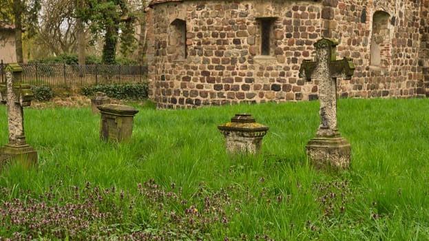 A historic stone cross in front of an old rustic church with small windows. Lively green grass in the foreground adds depth and contrast to the picture.