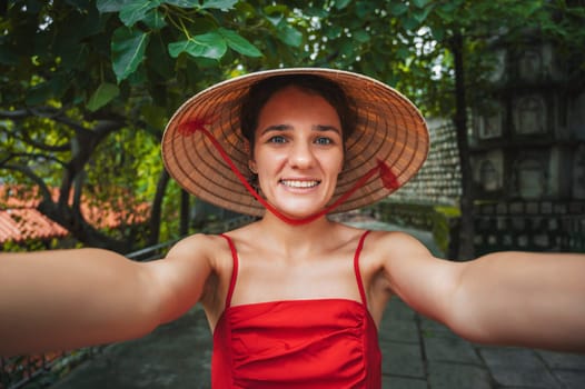 beautiful happy young female tourist traveler in a Vietnamese hat non La takes a selfie in garden in Vietnam. Tourism and travel in Asia