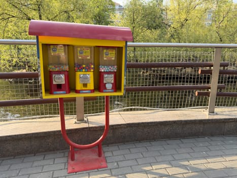 Bright vending machines with candy and chewing gum stand on city promenade by river. Cheerful colors contrast with greenery and calm water in background, evoking childhood memories