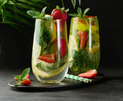 A refreshing drink in a glass with lemon slices, strawberries, fresh mint leaves, and sparkling water, served with lime and mint on a black plate