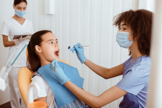 Dental procedure, drilling, tooth extraction. Millennial african american lady doctor in uniform, protective mask and gloves with tool, treat oral cavity of small hispanic kid in modern clinic