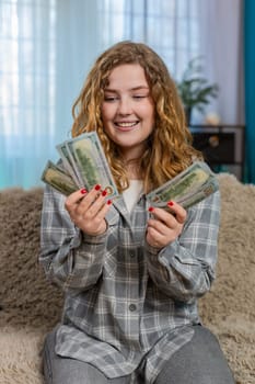 Young woman at home excitedly counting cash dollars in hands smiling and celebrating unexpected financial gain. Caucasian redhead girl on sofa happy about lottery win, salary or surprise money profit