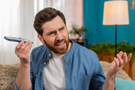 Young man at home carefully listening to a voice message on smartphone, tilting head and concentrating. Caucasian guy sitting on sofa, trying to hear clearly and focusing on important audio details.