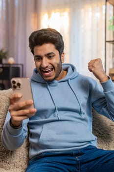 Surprised excited Indian young man holding smartphone, amazed by online game win or good news. Happy guy on sofa overjoyed by success, raises arms triumphantly, celebrating victory in living room.