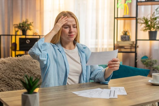 Young woman checks statement at home, eyes widening with alarm over unexpected expense. Girl sits at table sets paper aside, revealing tense realization of higher than normal cost of utility services