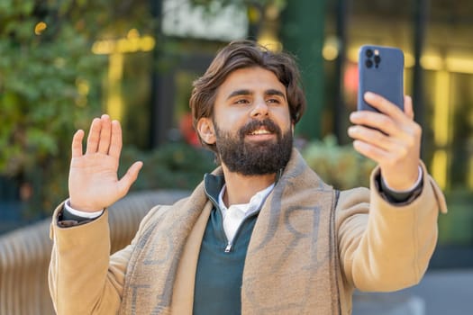 Indian young businessman taking selfie on smartphone, communicating video call online with subscribers, recording stories for social media vlog. Lebanese bearded guy blogger on downtown city street