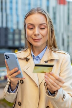 Caucasian mature businesswoman using credit card and smartphone while transferring money purchases online shopping order food delivery on internet outdoors. Girl against building on downtown street.