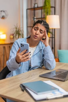 Upset displeased Indian woman freelancer using smartphone sitting at desk table in living room. Sad young girl touching head loses becoming surprised by sudden lottery results or bad news. Vertical.