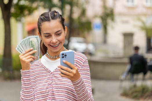Happy rich young woman with braces on teeth counting money dollar cash, use smartphone calculator app, plans to order gifts, food delivery online booking hotel room outdoors. Girl on urban city street