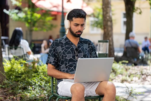 Indian bearded man in headphones working online distant job with laptop browsing website chatting sitting outdoors. Hispanic guy on city street looking at notebook screen send messages watching movies