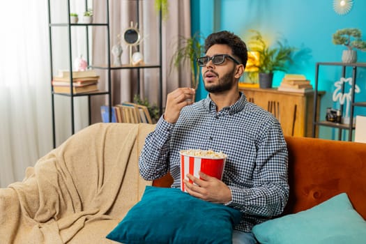 Indian man freelancer taking break from work wearing 3D glasses eating popcorn watching movie film content on TV. Smiling Hispanic guy sits at home couch sofa having snacks during weekends. Lifestyle