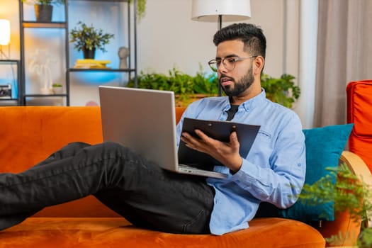 Happy young man freelancer work with documents on home sofa reading information business papers prepare financial report on laptop. Indian guy accountant businessman relaxing after finishing work.