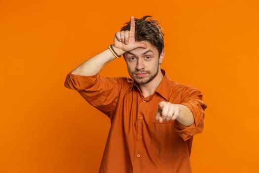 Caucasian man showing loser gesture and pointing on you, blaming accusing for unsuccess, expressing disrespect, mocking your failure, bullying abuse. Young guy isolated on orange background indoors