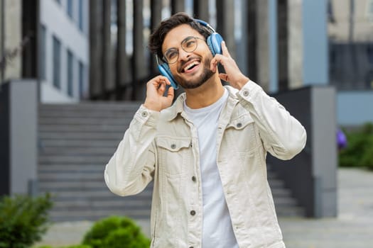 Happy relaxed young Indian man in wireless headphones dancing, listening favorite energetic disco music dancing outdoors. Arabian Hindu overjoyed guy standing in downtown city street. Business people