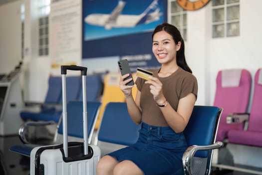 A cheerful young woman at the airport, displaying her credit card and phone, embodying the excitement of travel plans.