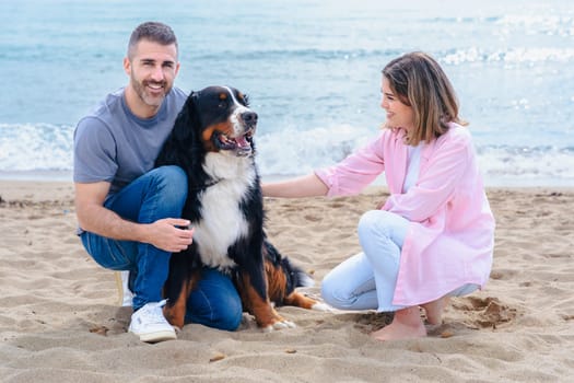 Portrait of a smiling caucasian couple with their large dog on the beach during vacation. Relaxed and joyful moment by the sea, with all three looking at the camera on a bright day.