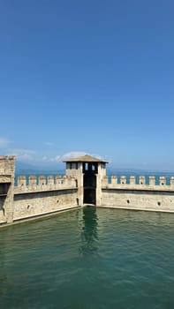 02.08.2024 Italy: Medieval Italian Castle Scaligero Walls Overlooking a Calm Blue Lake Garda Under a Clear Sky. A picturesque view of ancient 13th Century castle walls and tower surrounded by water, with scenic blue sky and serene lake Garda landscape in Sirmione, evoking a sense of history, tranquility, and natural beauty.