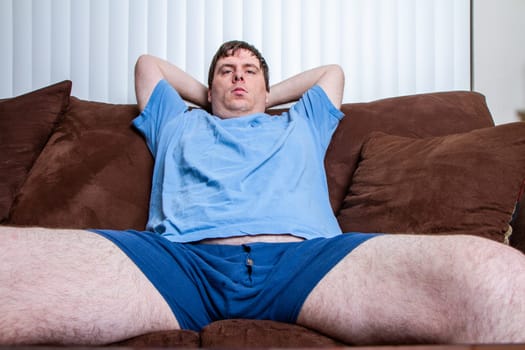 A man is sitting on a couch with his legs crossed and his arms behind his back. He is wearing a blue shirt and blue shorts