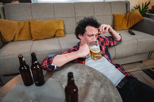 Young man drinking beer at home after party, lying on the floor by the sofa