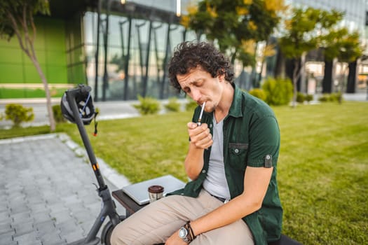 Man with curly hair sits on a park bench, lights a cigar and rests after riding an electric scooter