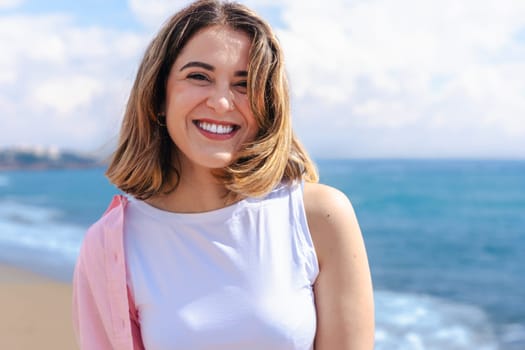 Portrait Happy caucasian woman on vacation standing on the beach, looking at the camera with a smile. Bright day, ocean in the background, clear sky and copy space on one side for design or text.