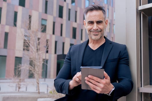 Confident businessman smiling while using a tablet outdoors in a modern urban setting.He wears a black suit and smartwatch. Concept of success, technology,digital lifestyle,and productivity.Copy space