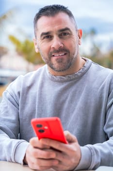 Caucasian man sitting at an outdoor cafe table, smiling at the camera while holding a red smartphone. Casual outfit and relaxed expression create a warm, approachable and modern tech vibe.vertical