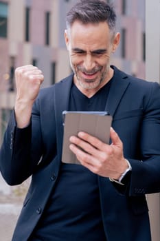 Confident businessman celebrates winning an online bet, holding a tablet and raising his fist. He smiles outdoors in a city setting. Concept of success, digital lifestyle, and modern finance. Vertical