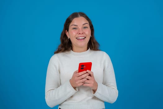 Cheerful young woman in white sweater looking at camera while holding a red smartphone in her hands, standing against a vivid blue background, representing connection, joy, and modern communication.