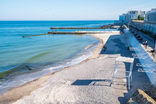 Embankment, sea sand beach in European city. People are walking in the distance. Sandy beach in midday sun. Concrete jetties go into calm sea.