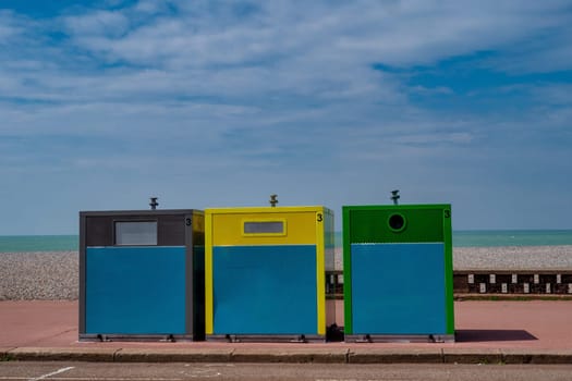 Three trash cans are lined up on the sidewalk, one of which is green. The other two are blue and yellow. The scene is set against a backdrop of a blue sky and a beach