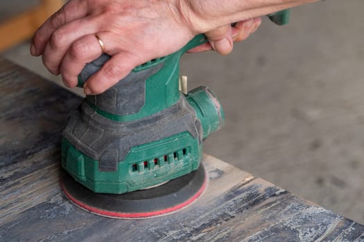 A man is using a sander to smooth out a surface. The sander is green and has a red handle. The man is wearing a ring on his finger