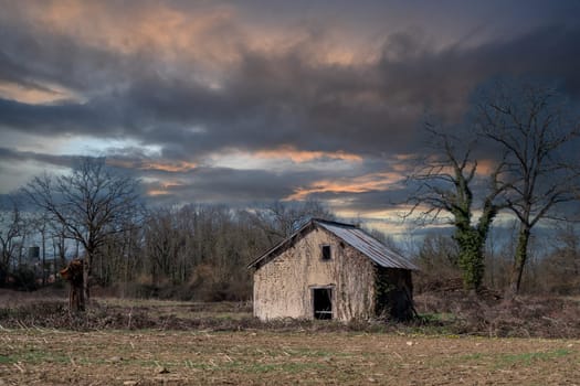 A small, old house sits in a field with a cloudy sky above. The house appears to be abandoned and the sky is overcast
