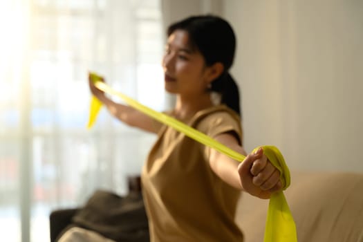 Asian woman using resistance band for arm exercises in a bright living room.