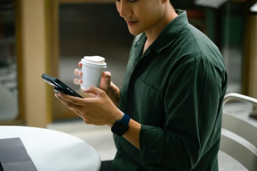 Man holding a smartphone and takeaway coffee cup while sitting at an outdoor cafe.