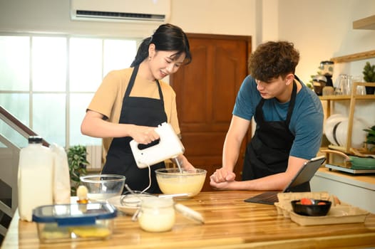 Smiling couple making pancake batter together using a hand mixer in a kitchen.