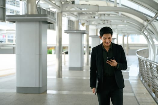 Confident executive walking through a modern city walkway and using smartphone.