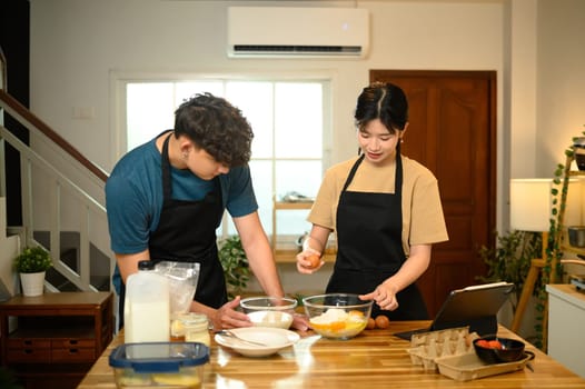 Young couple cracking egg into flour while preparing pancake batter together.
